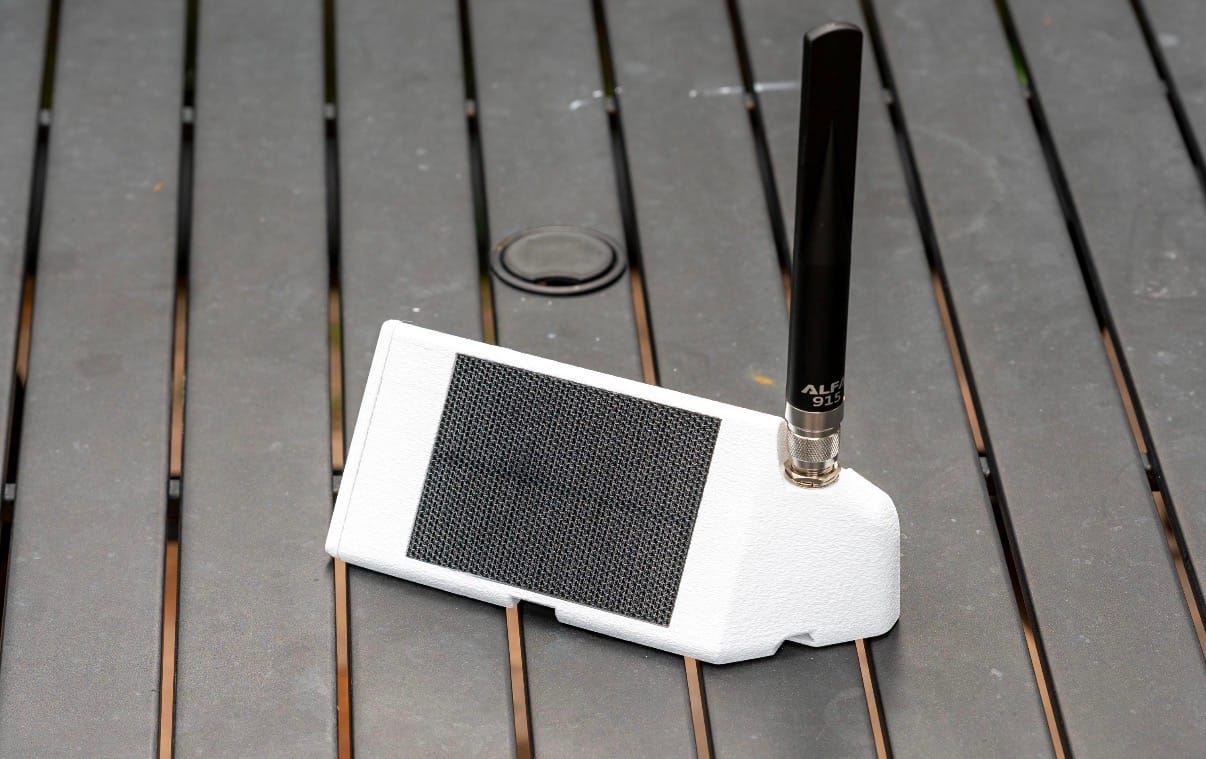White PeakMesh Magnet Mover meshcore node with ventilated top panel and 915MHz LoRa antenna, sitting on a wooden deck